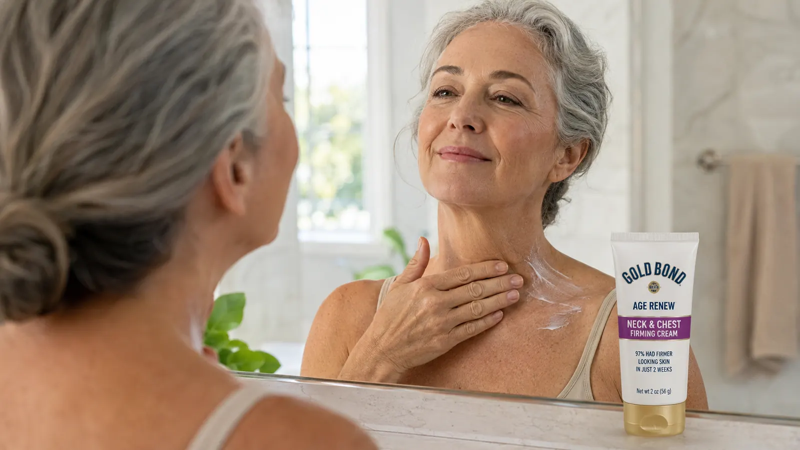 Woman in her sixties applying neck firming cream in front of a bathroom mirror, Gold Bond tube visible on the counter