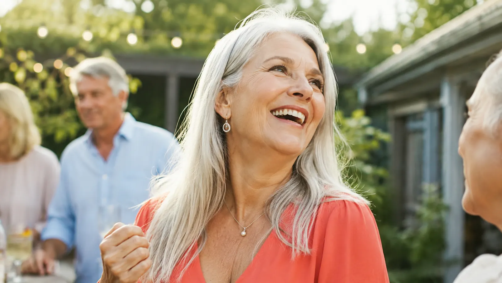 A woman in her sixties wearing a coral v-neck blouse laughs outdoors at a backyard gathering, chin up and relaxed, natural summer light.
