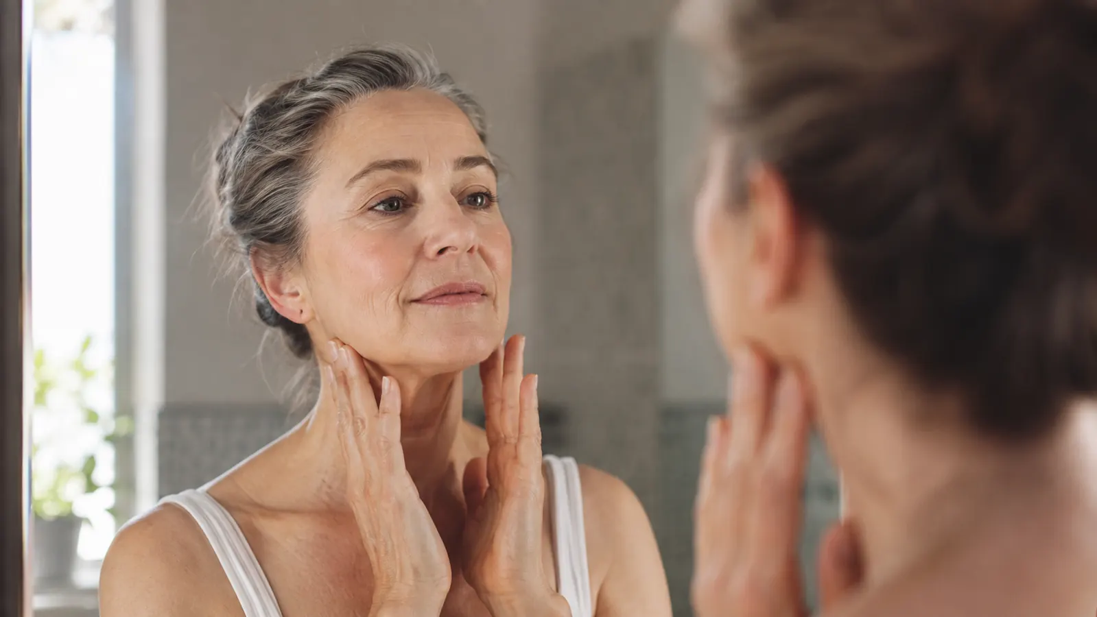 Woman in her sixties applying moisturizer to her neck and jawline in a bathroom mirror, morning light